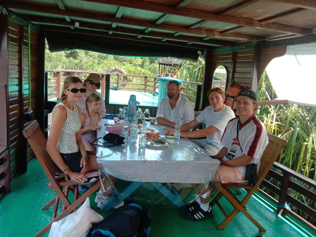 Tamu berkumpul untuk makan di dek bawah klotok dengan pemandangan tepi sungai hutan.