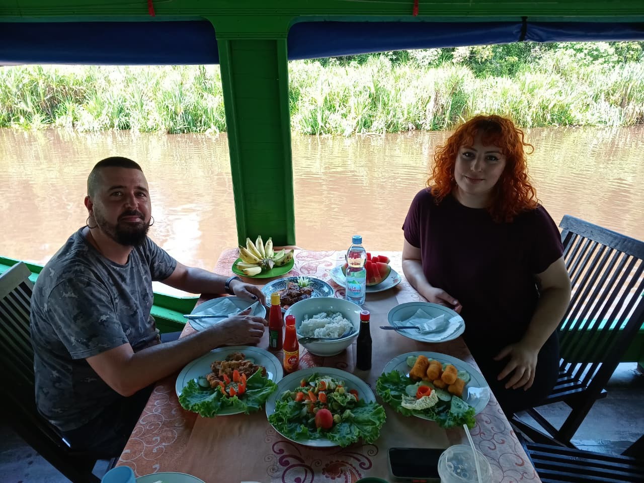 Pasangan menikmati makan siang masakan lokal segar di dek klotok di tepi sungai.